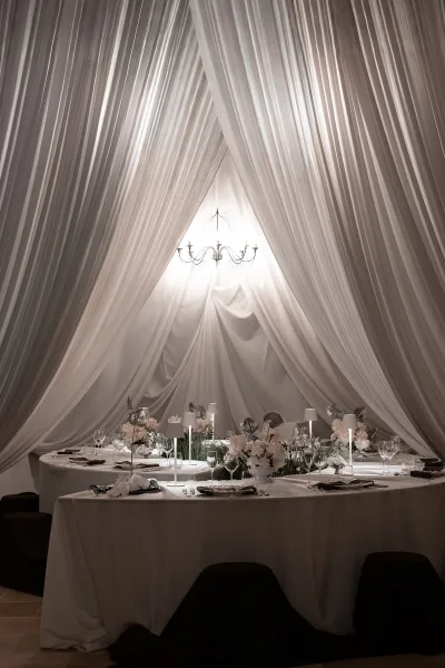 Reception tablescape with draped ceiling, chandelier glow, round white-linen tables, floral centerpieces, lamps, and black chairs in a tented hall