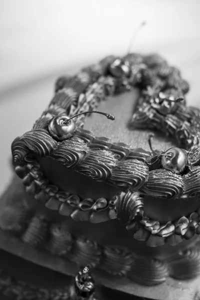 Wedding cake in black and white, heart-shaped with buttercream piping and icing ruffles, topped with cherries against a blurred backdrop