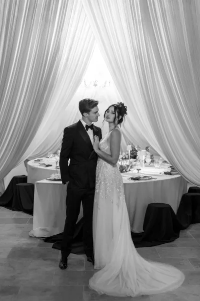 Couple portrait of bride and groom pose in black tie attire, standing by a banquet table under chandelier with draped fabric backdrop