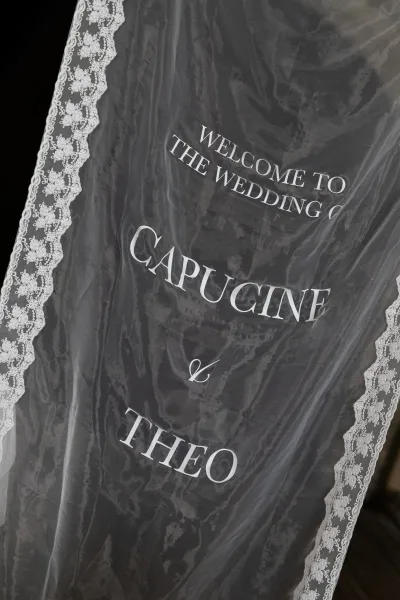 Wedding welcome sign on clear acrylic with white vinyl lettering and lace trim, standing against a dark backdrop at the entrance