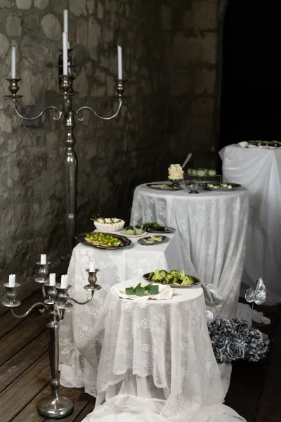 Wedding dessert table with white lace tablecloth and silver candelabra, set on round pedestals against a stone wall backdrop