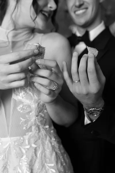 Wedding cake moment with newlyweds eating cake, hands showing wedding rings as blurred guests watch in an indoor reception setting