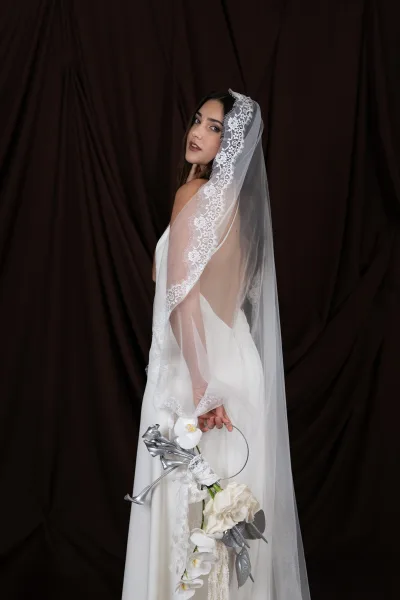 Bridal portrait of a bride looking over her shoulder in a white dress, holding an orchid bouquet behind her back against a dark backdrop