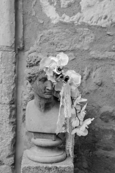 Wedding floral decor with white orchids draped over a stone bust statue, tied with lace ribbon, set against a rustic stone wall