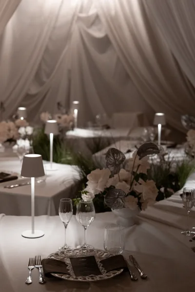Reception tablescape with wedding table decor featuring white florals, greenery runner, anthurium leaves, and cordless lamps under draped tent ceiling