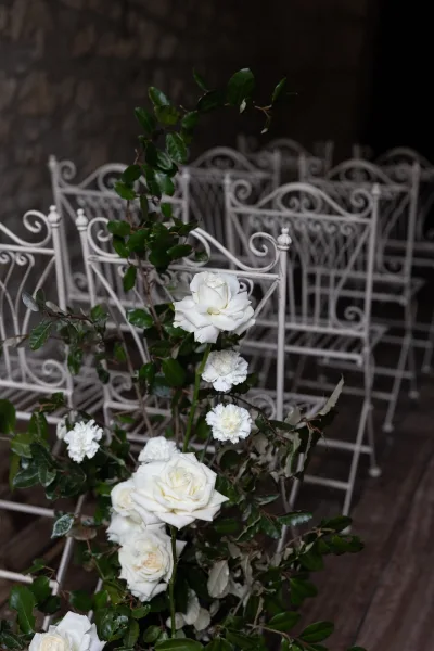 Ceremony aisle decor with white rose aisle flowers and greenery clusters beside white wrought iron chairs on a rustic wood floor