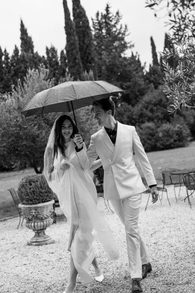 Couple portrait with a wedding umbrella as bride and groom walk on a gravel garden path, her veil trailing behind his white suit