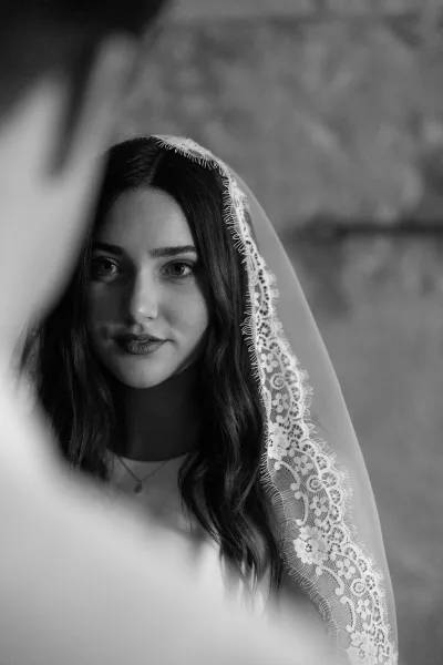 Bridal portrait in black and white of a bride in a lace veil and delicate necklace, posed by a stone wall with a blurred figure foreground