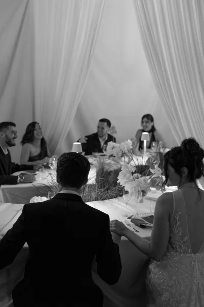 Reception toast moment as bride in wedding dress and groom in tuxedo listen hand in hand at a candlelit indoor table with white drapery backdrop