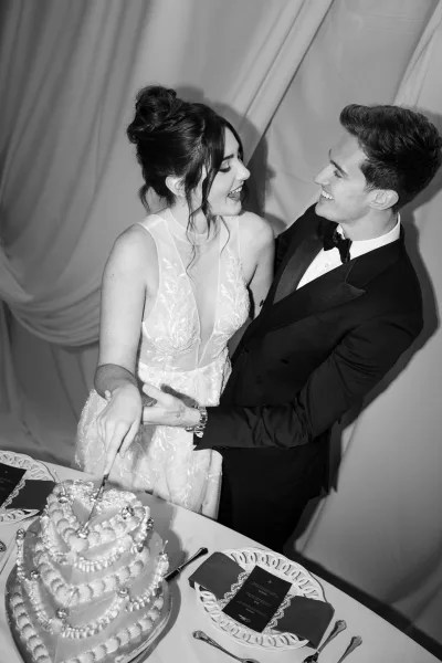 Wedding cake cutting as bride in lace gown and groom in tuxedo slice tiered cake with knife at reception table, draped curtain backdrop