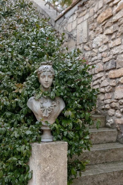 Garden statue with a stone bust planter on a pedestal, framed by leafy shrubs beside a stone wall and steps in an outdoor walkway