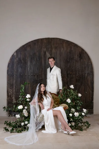 Couple portrait with bridal veil portrait as bride sits on a loveseat and groom stands behind, framed by arched wooden doors and florals