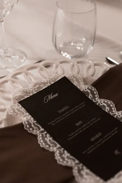 Wedding menu card in black calligraphy on a lace doily atop a white ceramic charger, with brown napkin, glassware, and silverware