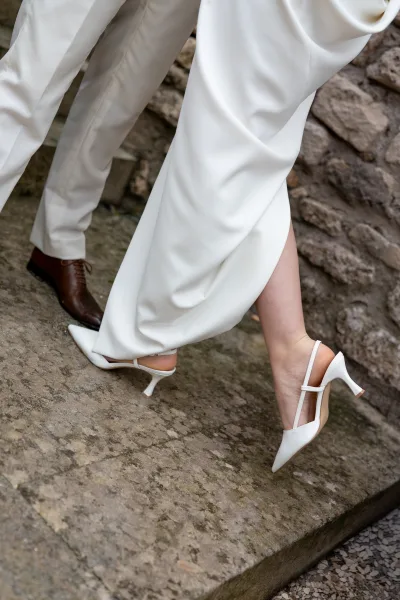 Wedding shoe detail of white bridal heels and groom’s brown dress shoes on stone steps, framed by dress hem and suit trousers