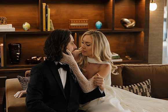 Couple portrait of bride and groom on sofa, bride touching groom’s face in lace gloves, with built-in bookshelf and warm wood interior behind.