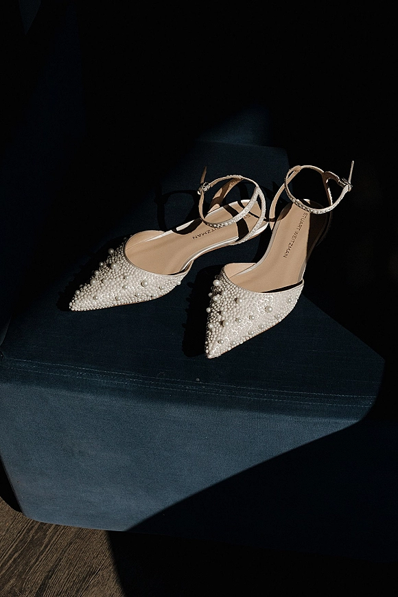 Bridal shoes with pearl wedding heels detail, pointed toe ankle-strap heels resting on a dark chair above a wooden floor