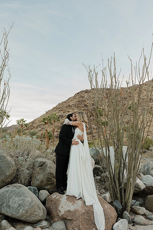 Wedding kiss portrait of bride and groom kissing, her long veil flowing over rocks, with desert mountains, ocotillo and palms behind