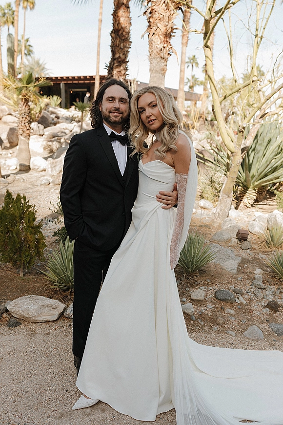 Couple portrait of bride in strapless gown with long veil and lace gloves beside groom in black tuxedo, in a desert garden with palms