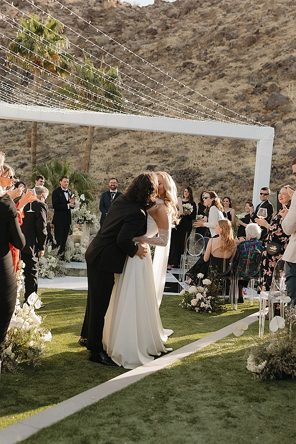 Wedding kiss as newlyweds share a ceremony kiss moment under string lights on an outdoor lawn, framed by palm trees and cheering guests