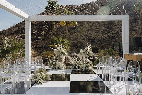 Ceremony setup with clear acrylic chairs and a black-and-white aisle runner leading to a white frame altar under string lights in desert mountains