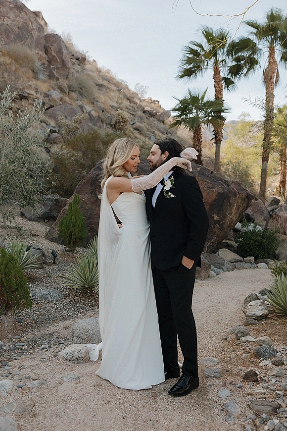 Couple portrait of a bride hugging her groom, veil and lace gloves flowing, on a gravel path with desert boulders and palm trees behind