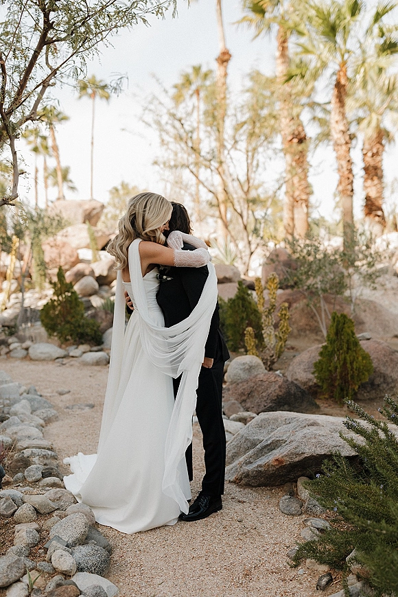 Wedding couple portrait of bride and groom hugging, her long veil trailing over a gravel path amid desert palms and boulders