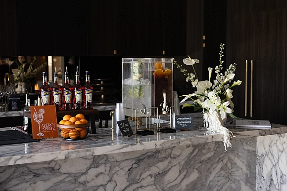 Wedding bar setup with Aperol spritz wedding bar sign, oranges, drink dispensers, ice, stacked cups, and white florals on a marble counter