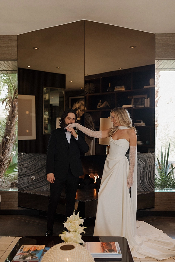 Couple portrait of groom kissing the bride’s lace-gloved hand in a modern living room with mirrored wall, fireplace, and windows to palms