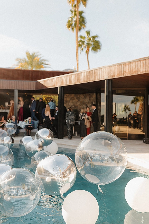 Pool party reception with clear balloons and white balloon decor floating in the pool beside a modern house patio with palm trees