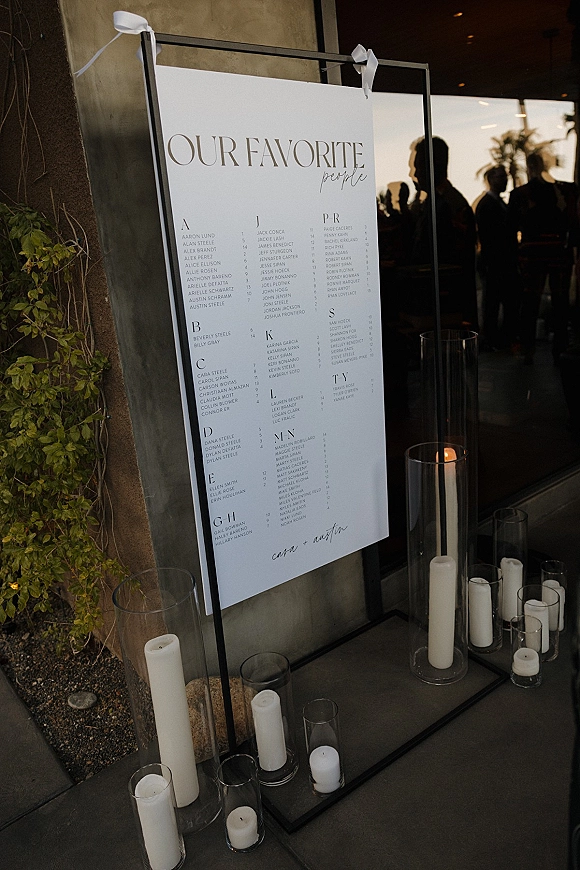 Seating chart sign in a black metal frame with a white alphabetical poster, ribbon bows, and candlelit hurricane vases by a window