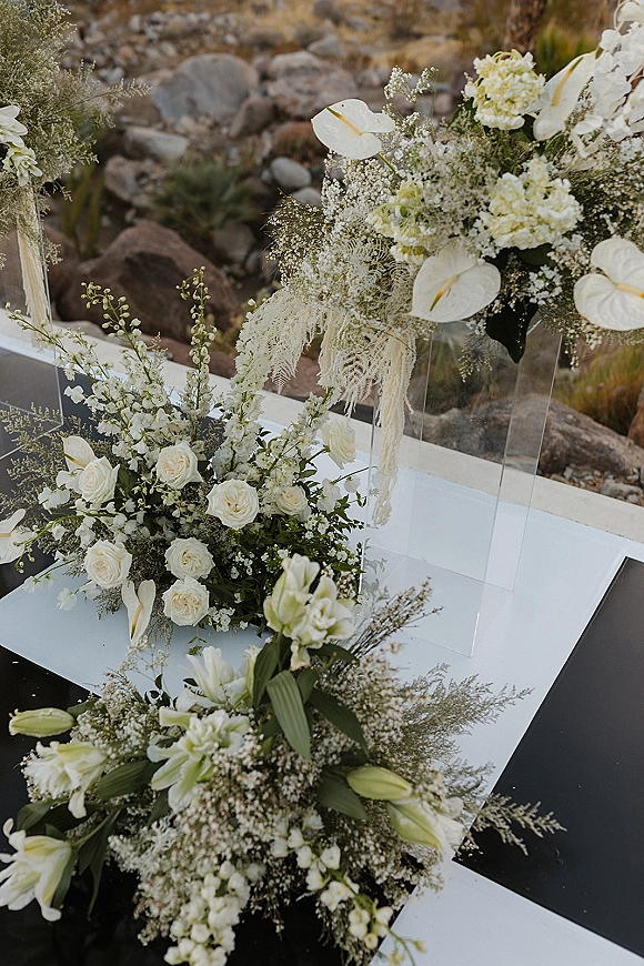 Wedding aisle decor with aisle floral arrangements of white roses, calla lilies, and greenery on clear acrylic stands in a rocky desert setting