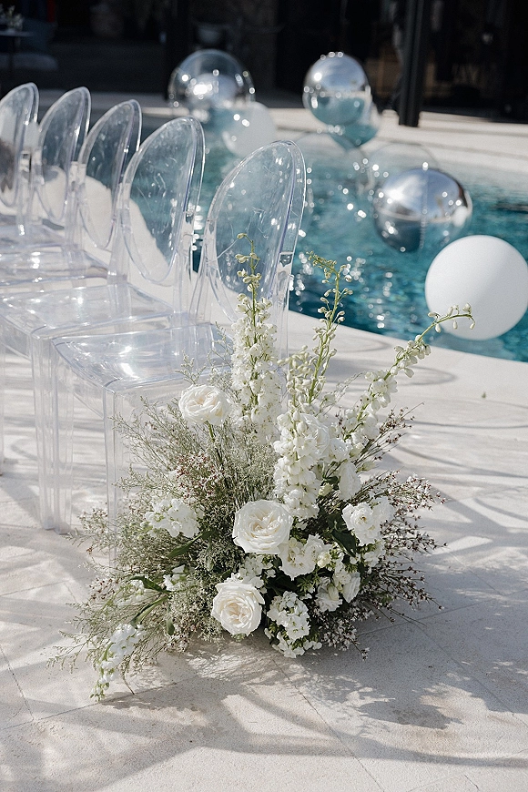 Ceremony aisle decor with clear acrylic chairs framing a white rose and greenery floral meadow, floating balloons by a pool deck backdrop