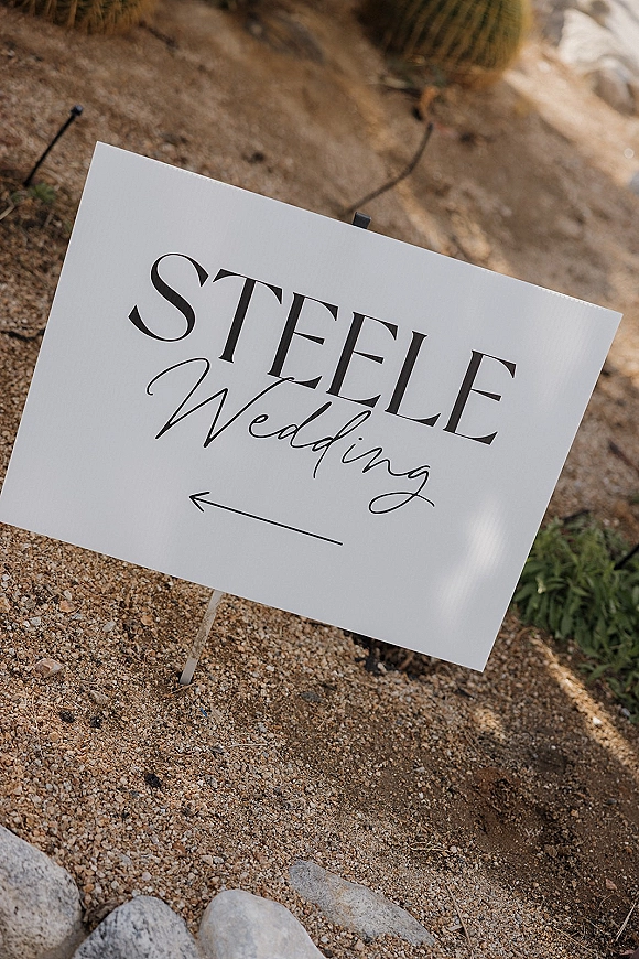 Wedding signage welcome wedding sign with black typography and arrow on a metal stake, set on gravel among desert plants and string lights