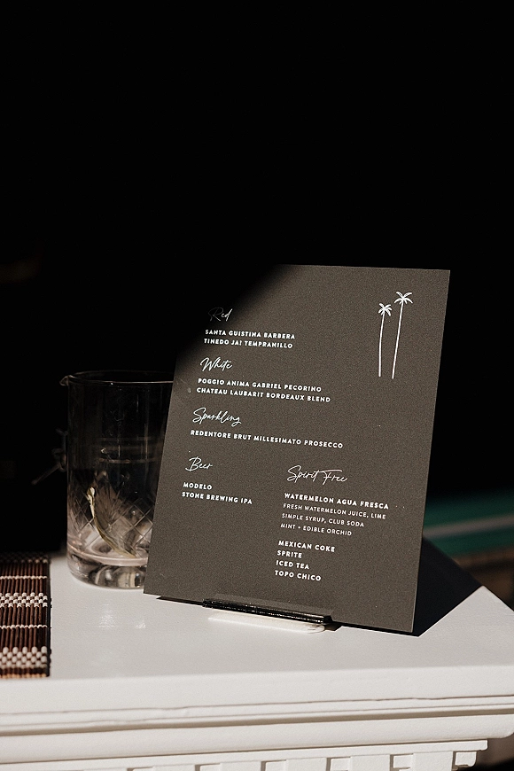 Wedding bar menu and signature cocktail menu card on a stand beside a drinking glass on a woven placemat at a dark bar counter