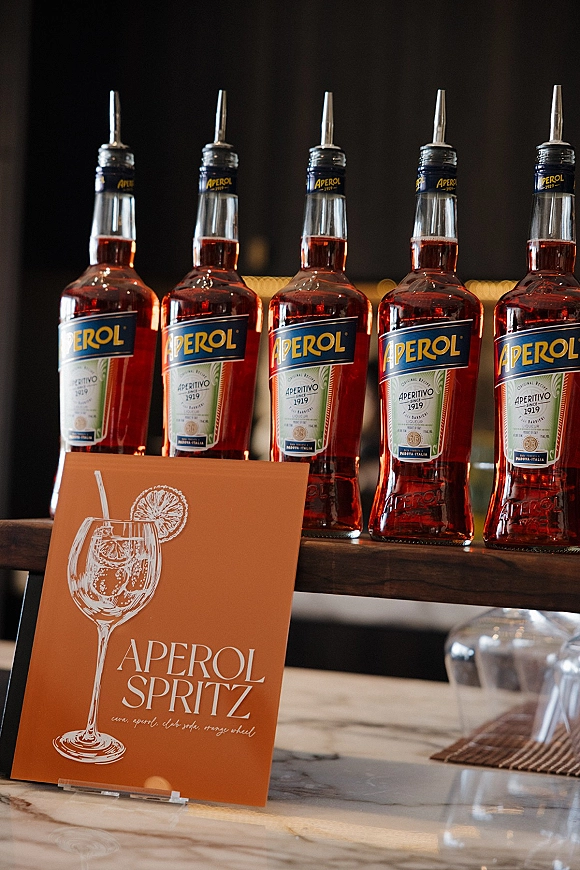Signature drink sign with a wedding signature cocktail sign in acrylic holder on marble bar, flanked by Aperol bottles and glassware