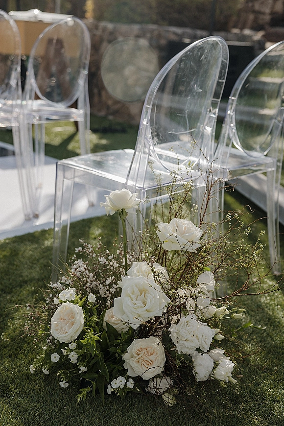 Ceremony aisle decor with outdoor ceremony chairs, a white aisle runner and white rose greenery florals on a sunlit lawn near a stone wall