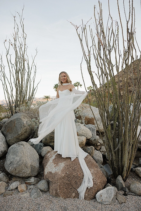 Bridal portrait of a bride in a white wedding dress with sheer draped sleeves and long train on boulders in a desert landscape with cactus and mountains