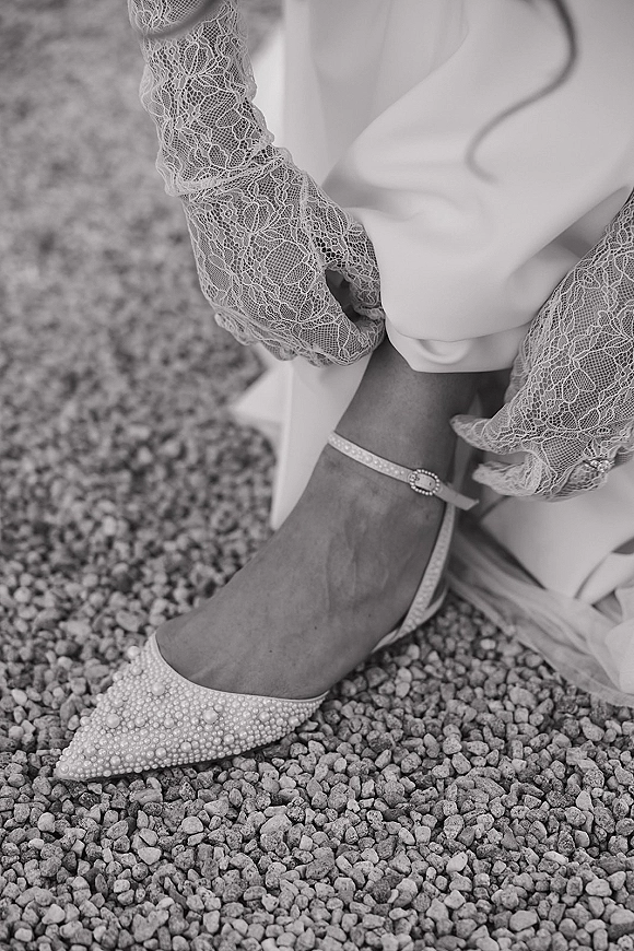 Bridal shoes with pearl wedding heels, pointed toe and ankle strap, shown with lace gloves and wedding dress hem on gravel ground