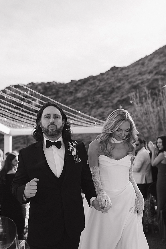 Wedding recessional as bride and groom walking hand in hand under string lights, guests cheering with a mountain landscape backdrop