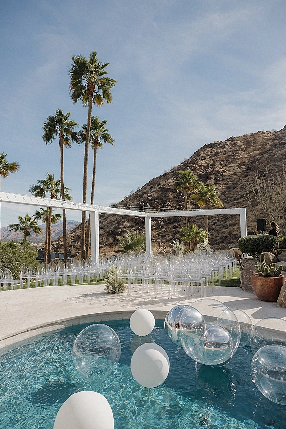 Ceremony setup with outdoor ceremony seating facing a white arch, clear acrylic chairs and aisle florals by a pool with balloon accents, palms and desert mountains