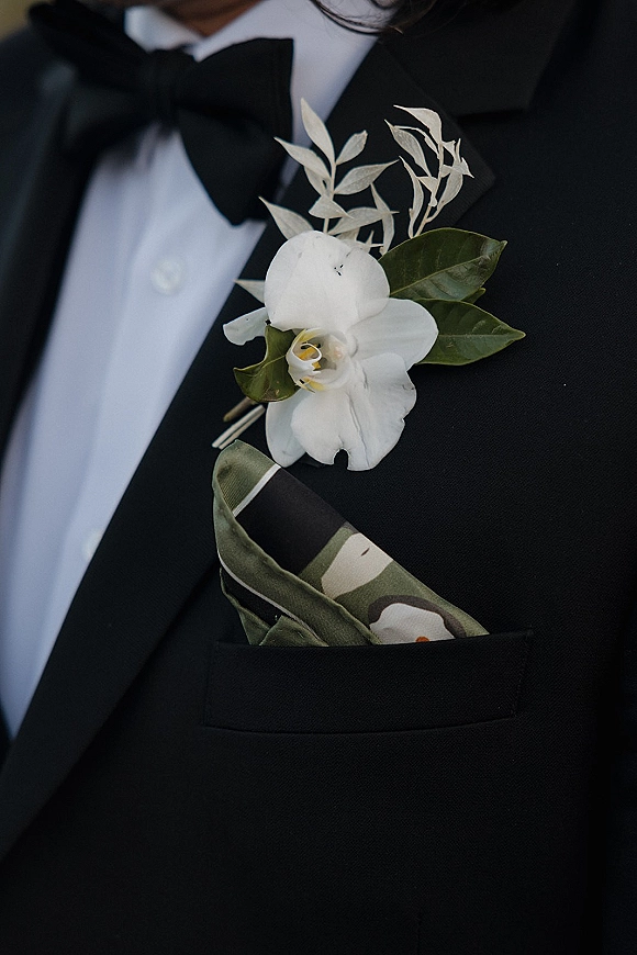 Wedding boutonniere with a white orchid and greenery pinned to a black tuxedo lapel, with bow tie and patterned pocket square visible