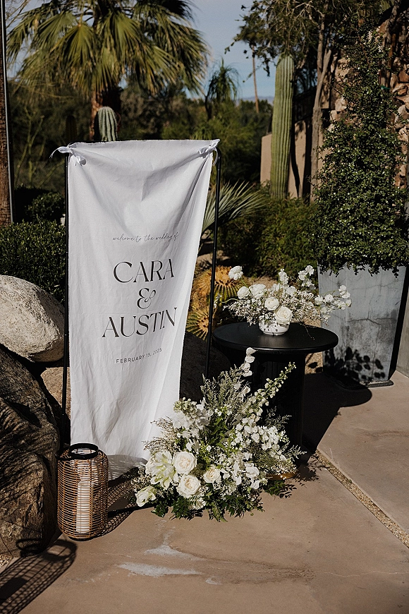 Wedding welcome sign on a black metal stand with white roses and baby’s breath on a pedestal table in a sunlit desert patio setting