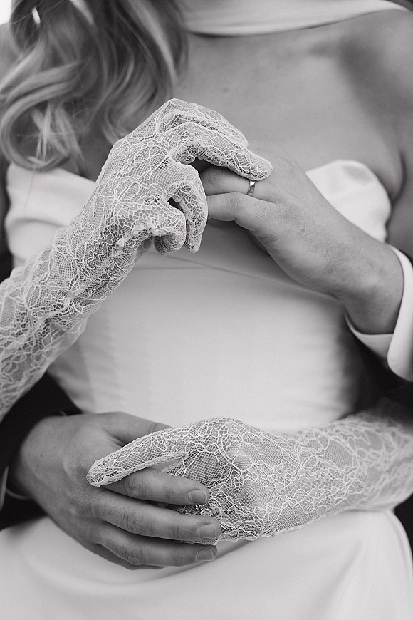 Wedding ring close-up showing wedding band photo on bride’s lace bridal glove as the couple embraces, groom’s cuff and necklace visible