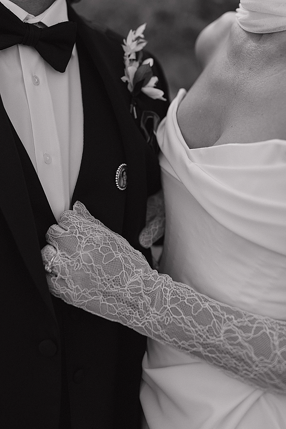 Couple portrait in a black and white wedding portrait, bride holding groom’s arm in tuxedo with boutonniere against greenery backdrop