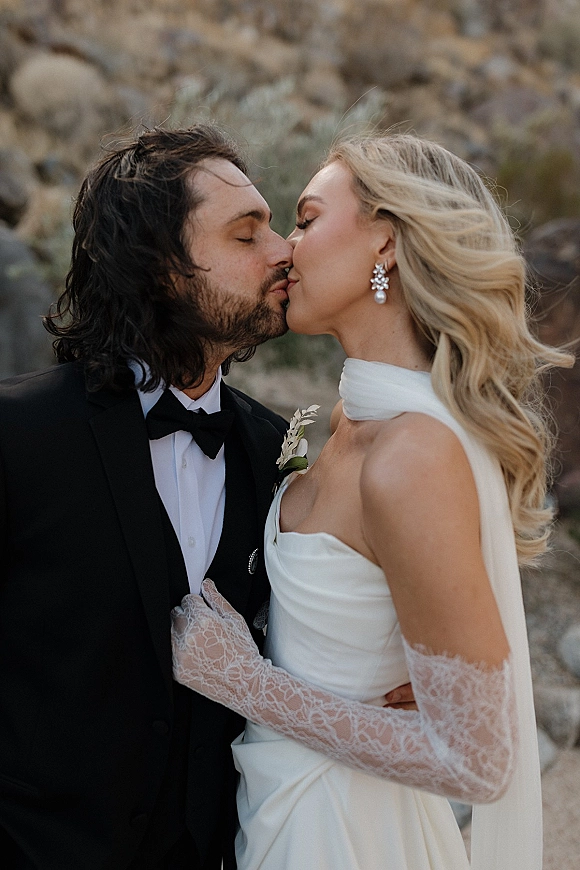 Wedding kiss portrait of bride and groom kissing, her strapless satin dress and veil visible against a rocky desert boulder backdrop