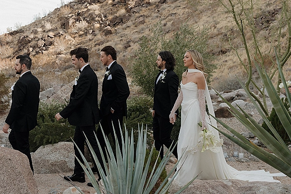 Wedding party portrait of bride and groomsmen walking along a gravel path, bride in veil with white bouquet against rocky desert hillside