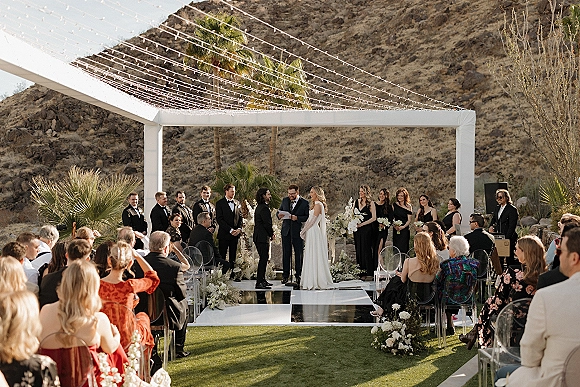 Wedding ceremony with a modern wedding arch under string lights, bridal party in tuxedos and black dresses, desert mountains behind