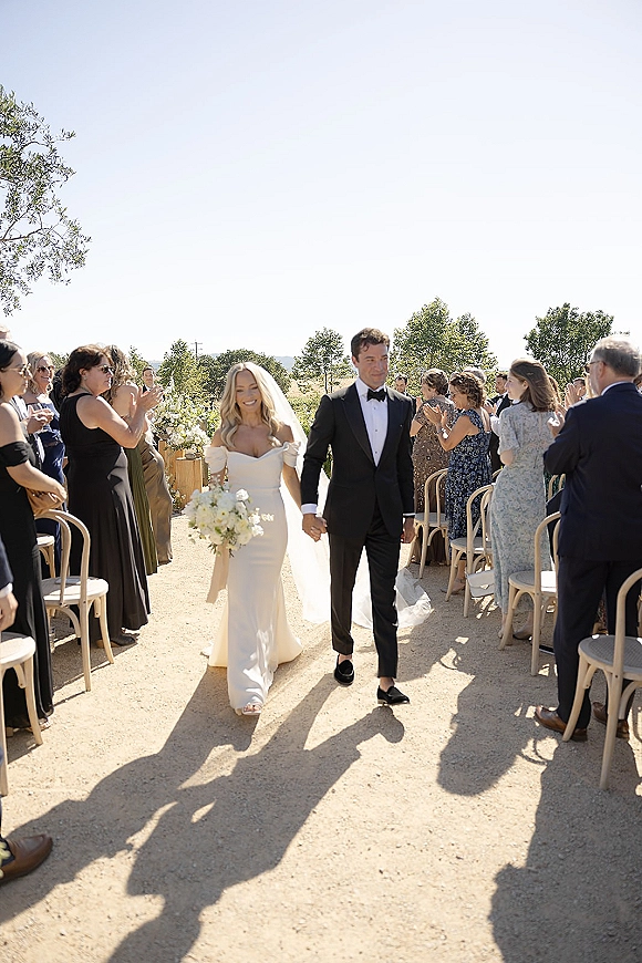 Wedding recessional as bride and groom walk hand in hand down a gravel aisle, bouquet and veil flowing, guests cheering under blue sky