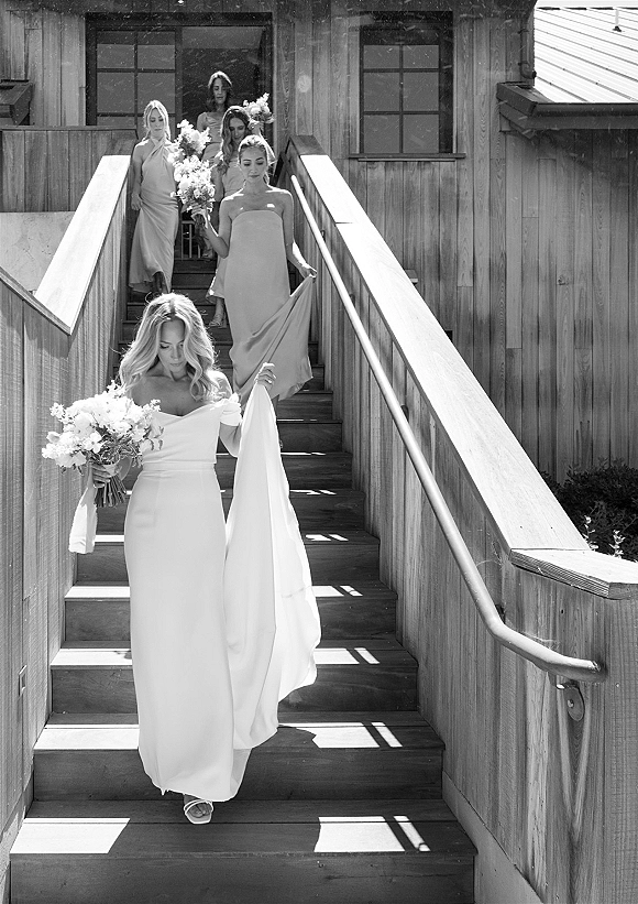 Bride and bridesmaids walking down stairs holding white bouquets, the bride in an off-the-shoulder gown on a sunlit wooden staircase