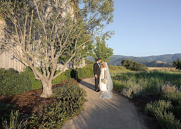 Couple portrait of bride and groom kissing, holding a white bouquet as her veil blows, on a gravel path by a barn under blue sky